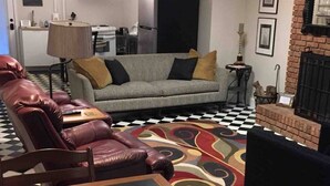 A living room with a sofa, a leather chair, a patterned rug, a fireplace, and a kitchen area in the background. - Cozy Suite on Cedars 1300sq ft (ONE LEVEL) (Hickory)