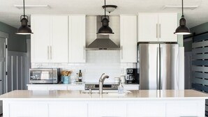 A modern kitchen with white cabinets, stainless steel appliances, and three bar stools. - Bear Inn OBX - 4X4 Carova Beach, Oceanfront, Ocean View, Hot Tub, Pets OK (4X4 - Carova Beach)