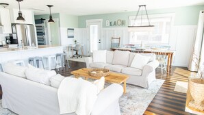 A modern living room with a white sofa, a wooden coffee table, and a kitchen area with white cabinets and a refrigerator. - Bear Inn OBX - 4X4 Carova Beach, Oceanfront, Ocean View, Hot Tub, Pets OK (4X4 - Carova Beach)