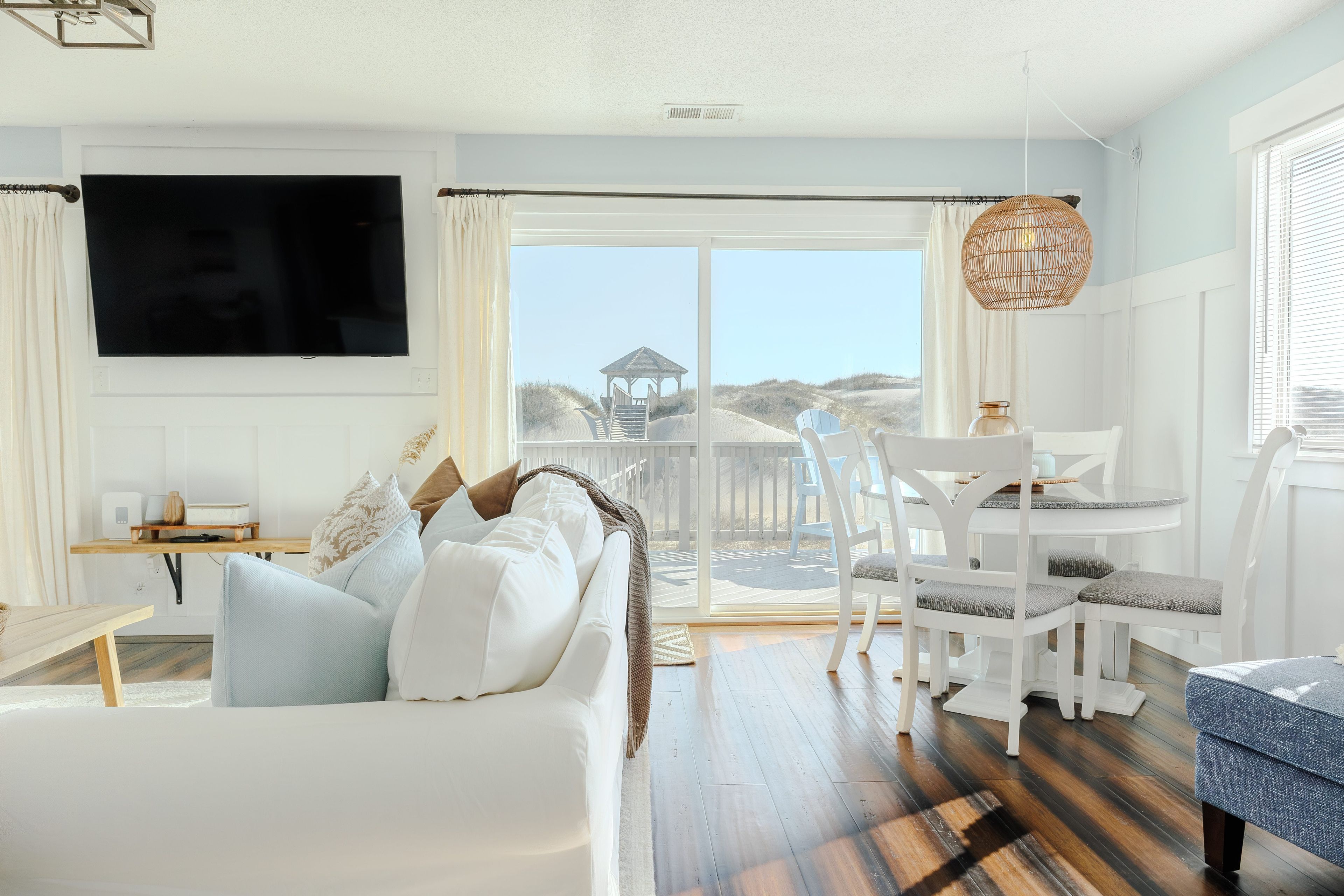 Un salon moderne doté d’un grand téléviseur, d’un canapé avec des coussins, d’un coin repas avec des chaises et d’une vue sur la plage, visible à travers la porte vitrée.