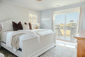 Une chambre avec un grand lit, une fenêtre avec des stores, une vue sur la plage et une terrasse en bois à l’extérieur.