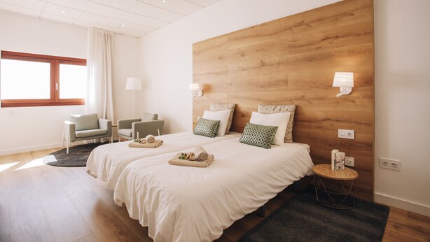 Quarto de hotel moderno com duas camas, cabeceira de madeira, uma mesa redonda pequena, uma cadeira e uma janela com cortinas.
