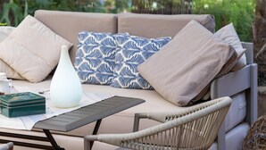 A cozy outdoor seating area with a cushioned sofa, a wicker chair, and a table with a white vase and a green box.