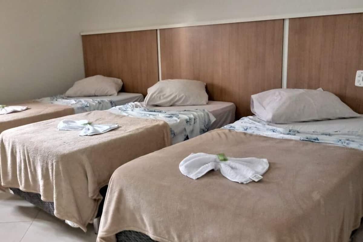 Una habitación de hotel con tres camas individuales, cada una con una colcha color beige y una almohada. La habitación tiene un cabecero de madera y piso de baldosas.