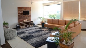 A modern living room with a sofa, a TV mounted on a wooden wall, a ceiling fan, and large windows with blinds. - Hotel Canaã Express (Jarinu)