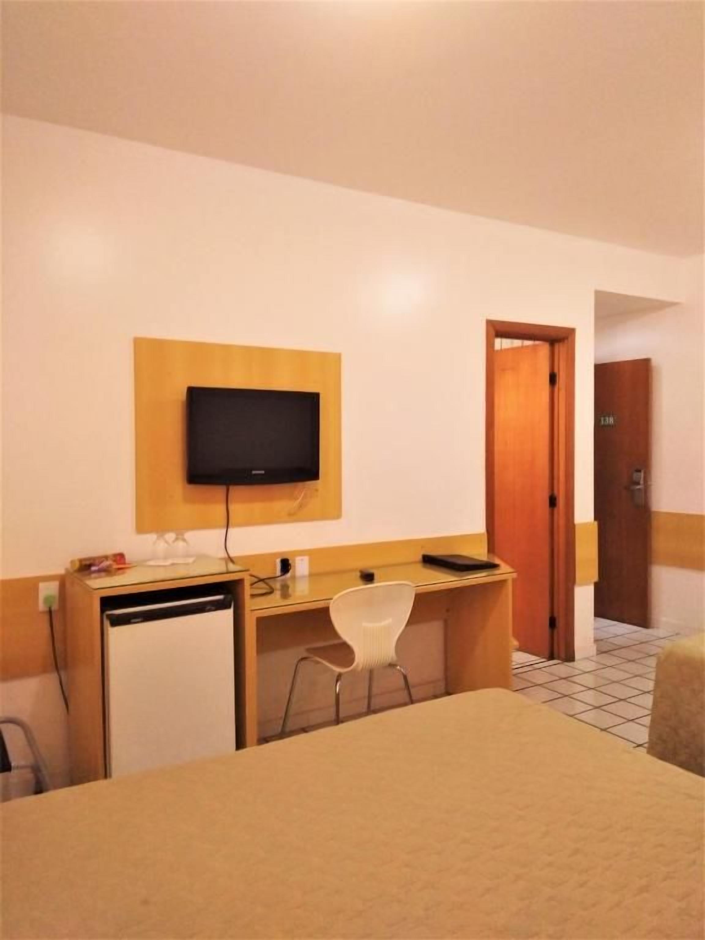 A hotel room with a bed, a desk, a chair, a television mounted on the wall, and a door.