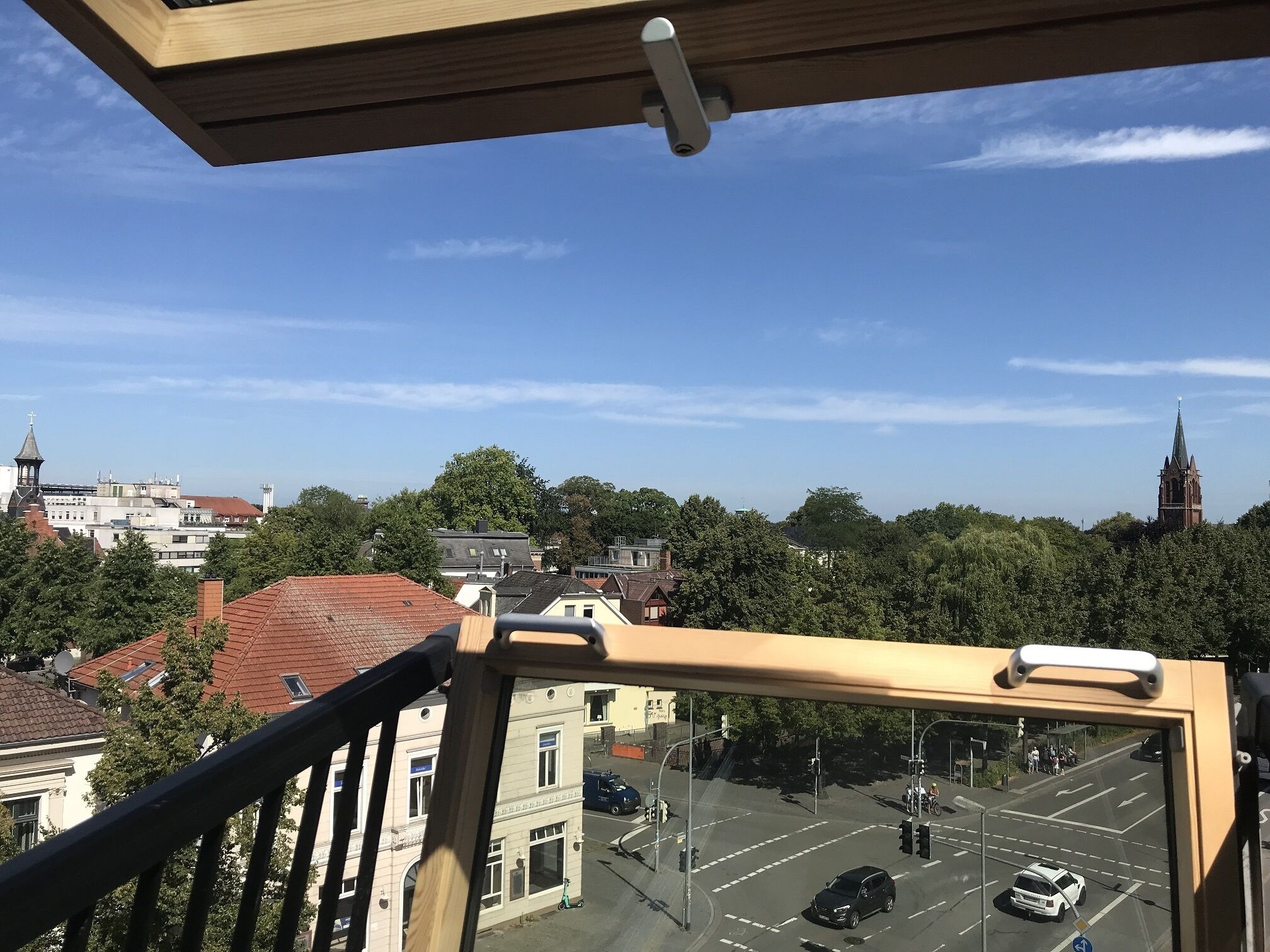Blick vom Balkon auf eine Stadtlandschaft mit Gebäuden, Bäumen und einem Kirchturm.