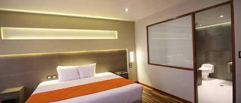 Habitación de hotel moderna con una cama grande, una mesita de noche, un baño con inodoro y lavamanos, y un armario con puerta corredera.