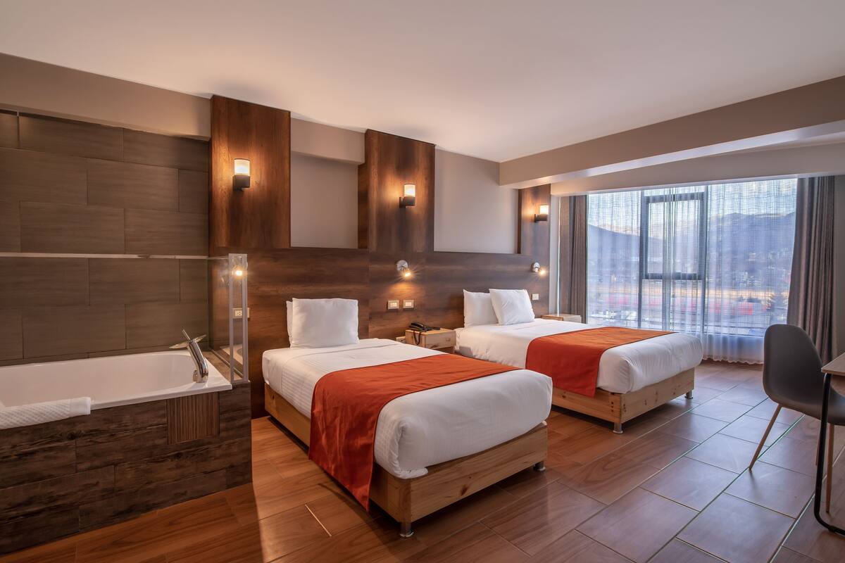 A modern hotel room with two beds, a large window, a bathtub, and a chair.