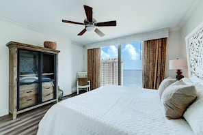 A hotel room with a large bed, a ceiling fan, a view of the ocean, and a wardrobe with a decorative mirror.