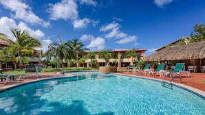 A swimming pool with lounge chairs and palm trees, surrounded by buildings with thatched roofs. - Cozy and spotless 6 guest apartment-great location only 50 steps to Eagle Beach! (Eagle Beach)
