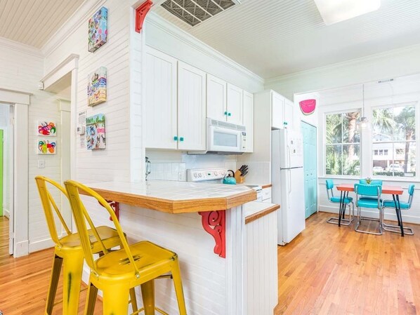 A kitchen with white cabinets, a microwave, and a refrigerator. There are yellow chairs and a table with blue chairs. - Coastal Soul Cottage | Pet Friendly, Fenced Yard (Tybee Island)