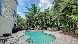 Una piscina con un patio de ladrillo, rodeada de exuberante vegetación tropical y un edificio con una parrilla cubierta.