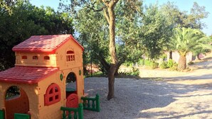 Una casita de juegos infantil con techo rojo y detalles verdes, rodeada de árboles y grava.