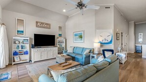 A spacious living room with a large sofa, a television, and a ceiling fan. There is a bookshelf and a small table with a lamp.