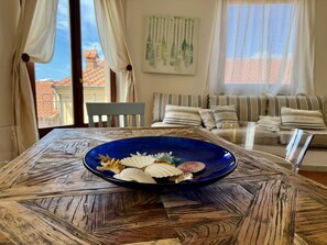 A dining table with a blue plate of food, a wooden table with a geometric pattern, a striped sofa, and a view of buildings through a window.
