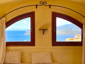 A room with two arched windows offering a view of the sea and a coastal town.