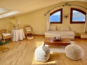 Um quarto aconchegante com um sofá, duas cadeiras de vime, uma mesinha com abajur, uma mesinha de centro e vista para o mar.