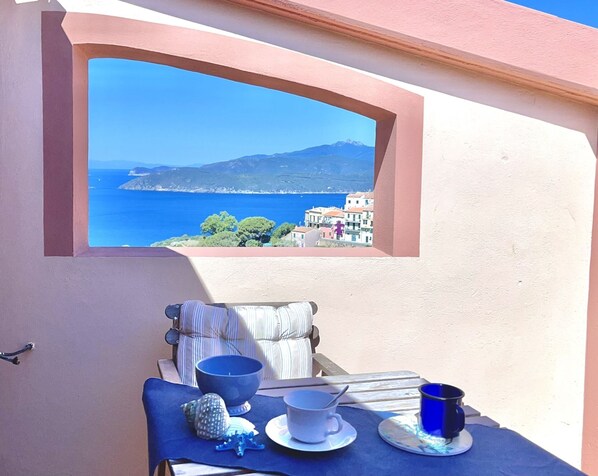 A balcony with a table set for two, a view of the sea, and a building with a pink facade.