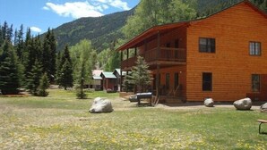 A two-story wooden lodge with a balcony, surrounded by a grassy area with rocks and trees, set against a mountainous backdrop. - Soltow's Place - Wonderful Duplex with Room For Everyone (Lake City)