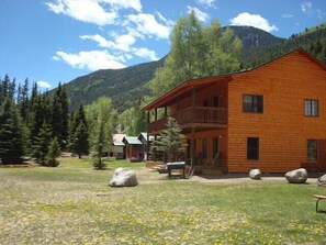 A two-story wooden lodge with a balcony, surrounded by a grassy area with rocks and trees, set against a mountainous backdrop.