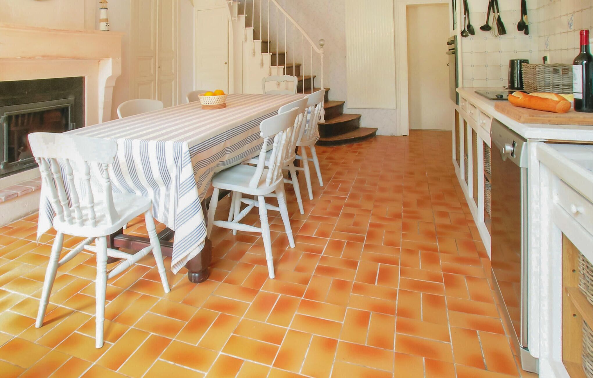 A dining area with a striped tablecloth, white chairs, a basket of oranges, and a fireplace.