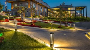 A modern building with a flat roof, large windows, and a landscaped garden with red flowers and trees. - Menalo Hotel Premium Istanbul Airport (Arnavutköy)