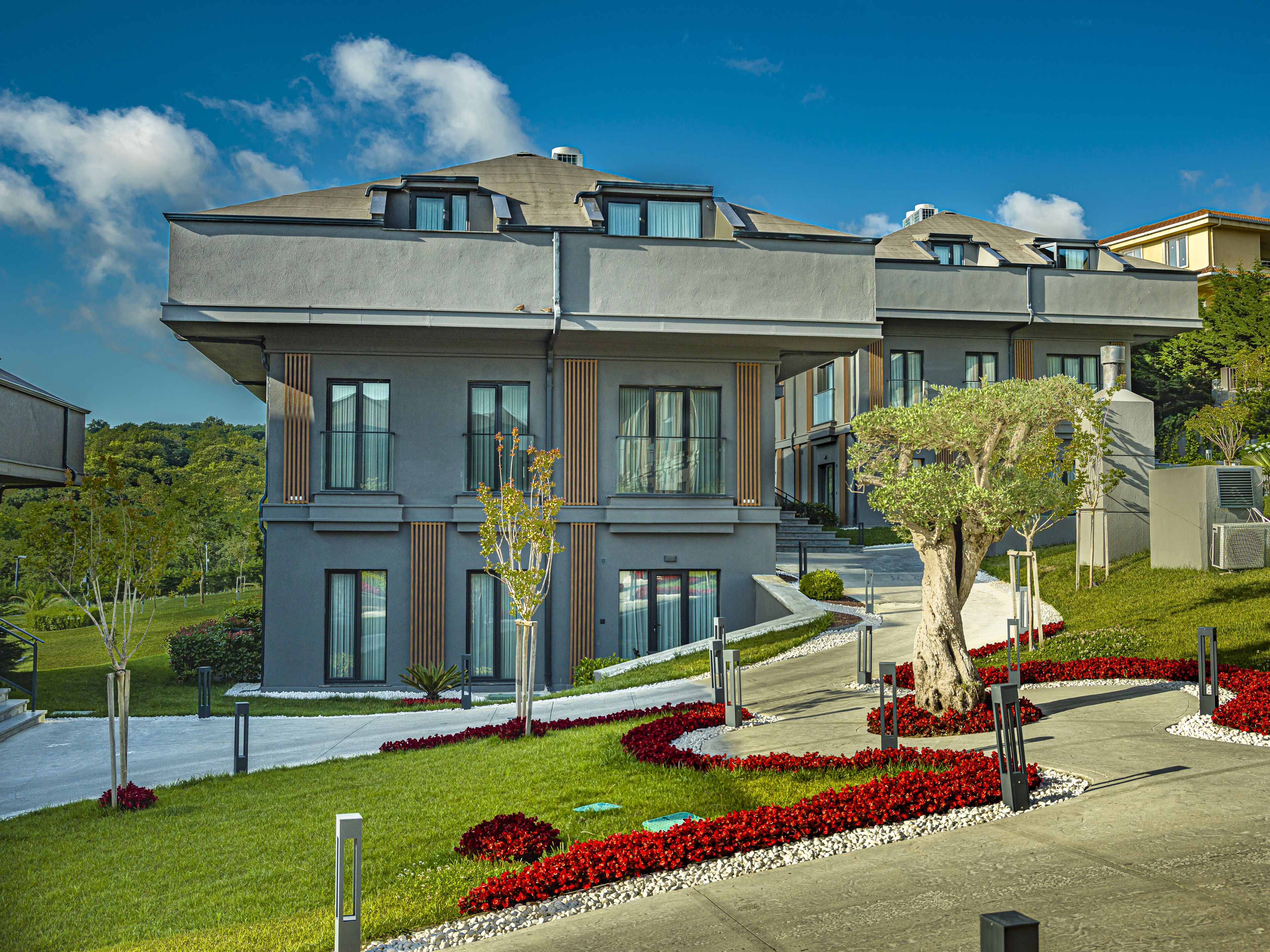 A modern multi-story residential building with a flat roof, large windows, and a landscaped garden with red flowers and a tree.