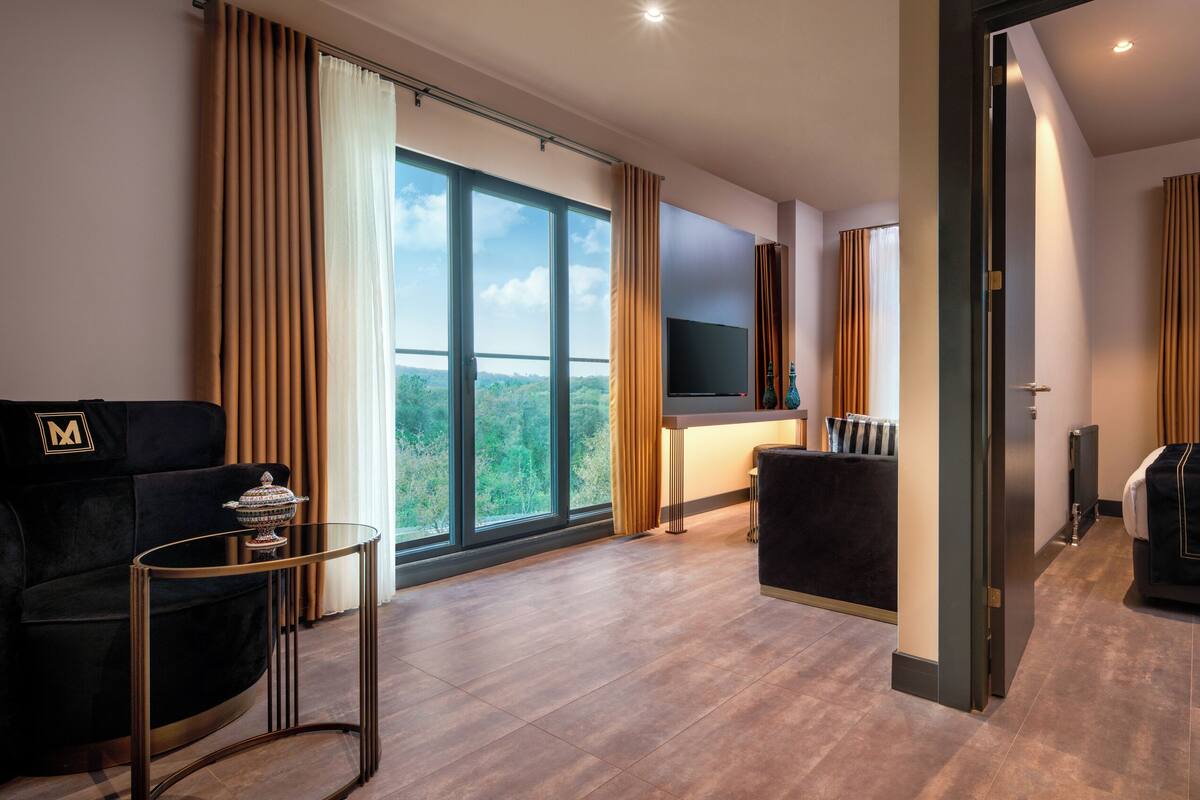 A modern hotel room with a large window, a sofa, a TV, and a small table.
