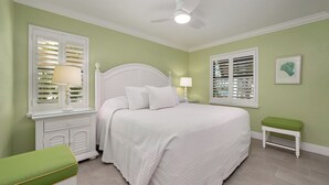 Une chambre avec un lit blanc, deux tables de chevet avec des lampes, un banc vert et un ventilateur de plafond.