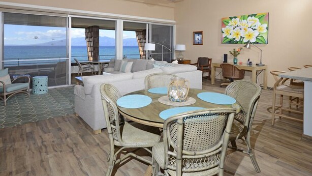 A room with a large window offering a view of the sea, a dining area with wicker chairs and a round table, and a sofa set.