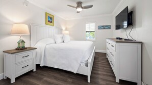 Een slaapkamer met een bed, nachtkastje, lamp, dressoir, plafondventilator, televisie en een raam met jaloezieën.