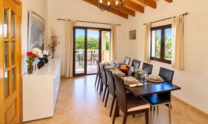 Uma sala de jantar com uma mesa comprida posta para seis, um balcão de cozinha com um vaso de flores e vista para o exterior através de portas de vidro.