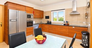 Een moderne keuken met houten kastjes, roestvrijstalen apparatuur, een eettafel met een schaal fruit, en uitzicht op groen buiten.