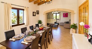 Een eetgedeelte met een lange tafel gedekt voor een maaltijd, een woonkamer met een bank en een eettafel, en een keuken met een koelkast.