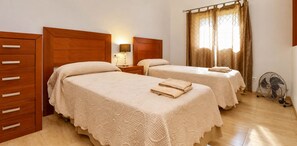 Quarto de hotel com duas camas, uma cômoda de madeira, uma mesa de cabeceira com abajur e um ventilador.