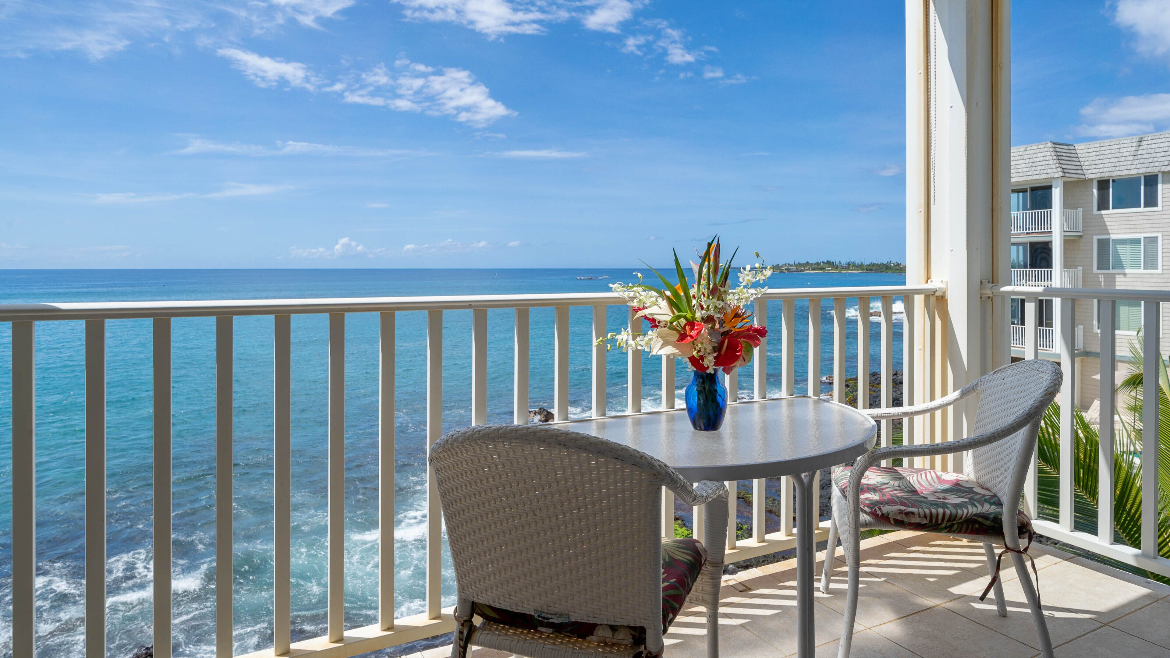 A balcony with a view of the ocean, a table with a vase of flowers, and two wicker chairs.