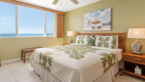 Habitación de hotel con una cama grande, vista al océano, un cuadro de una playa y una mesita de noche con lámpara.