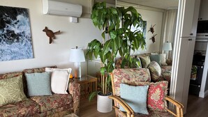 A living room with a sofa, armchair, and a glass coffee table. There is a large potted plant, a wall-mounted air conditioner, and a kitchen area visible in the background. - REDUCED RATES with A/C! 113 Darling little ground floor 1 bed, 1 bath (Lahaina)