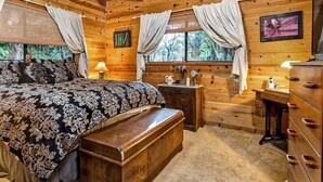 A cozy log cabin bedroom with a large bed, a dresser, a nightstand, and a window with curtains.