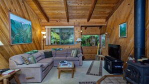 A cozy cabin interior with a sectional sofa, a television, a fireplace, and large windows offering a view of the surrounding forest.