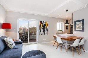 A modern living room with a sofa, a dining area with a wooden table and chairs, and a large window with a view of the outside. - Bright Visconde (Cascais)