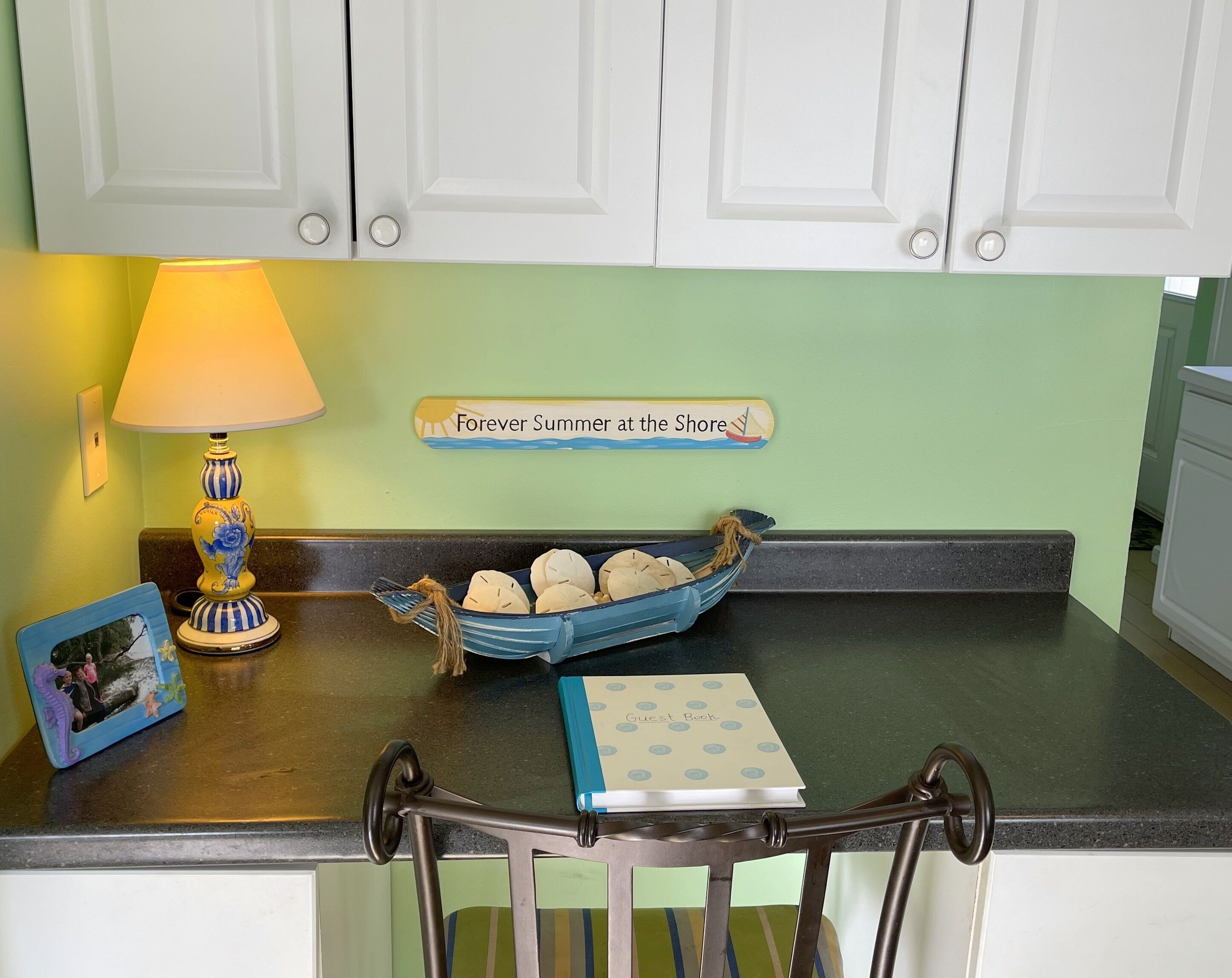 A kitchen counter with a decorative lamp, a guest book, a photo frame, and a sign that reads 'Forever Summer at the Shore.'