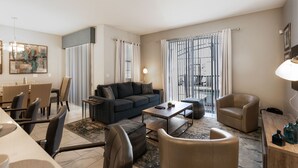 A modern living room with a sofa, coffee table, and chairs. There is a dining area with a table and chairs. The room has large windows with curtains and a view of a pool.