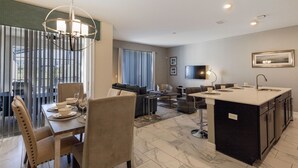 A modern kitchen with a central island, dining area with chairs and table, and a living area with sofas and a TV.