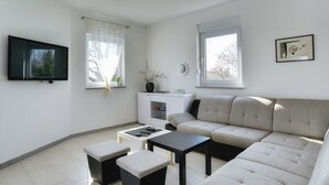 A modern living room with a sofa, two ottomans, a coffee table, a TV, a small cabinet, and a window with a view of trees. - Sunny villa with private pool (Majmajola)
