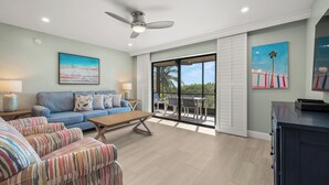 A spacious living room with a blue sofa, striped armchair, and a coffee table. A ceiling fan and artwork are visible. - Newly Renovated Beach Front 2 bed, 2 bath at Sanibel Moorings #1121 (Sanibel)