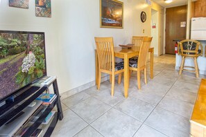 Un coin repas avec des chaises en bois et une table, un téléviseur posé sur un meuble, et un espace cuisine équipé d’un réfrigérateur.
