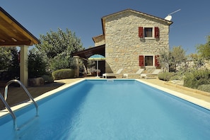 Una piscina con un edificio in pietra sullo sfondo, una zona coperta con tavolo e sedie, e un cielo azzurro intenso.