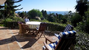 Une terrasse extérieure avec une table ronde et des chaises, entourée d’une végétation luxuriante et offrant une vue sur la mer.